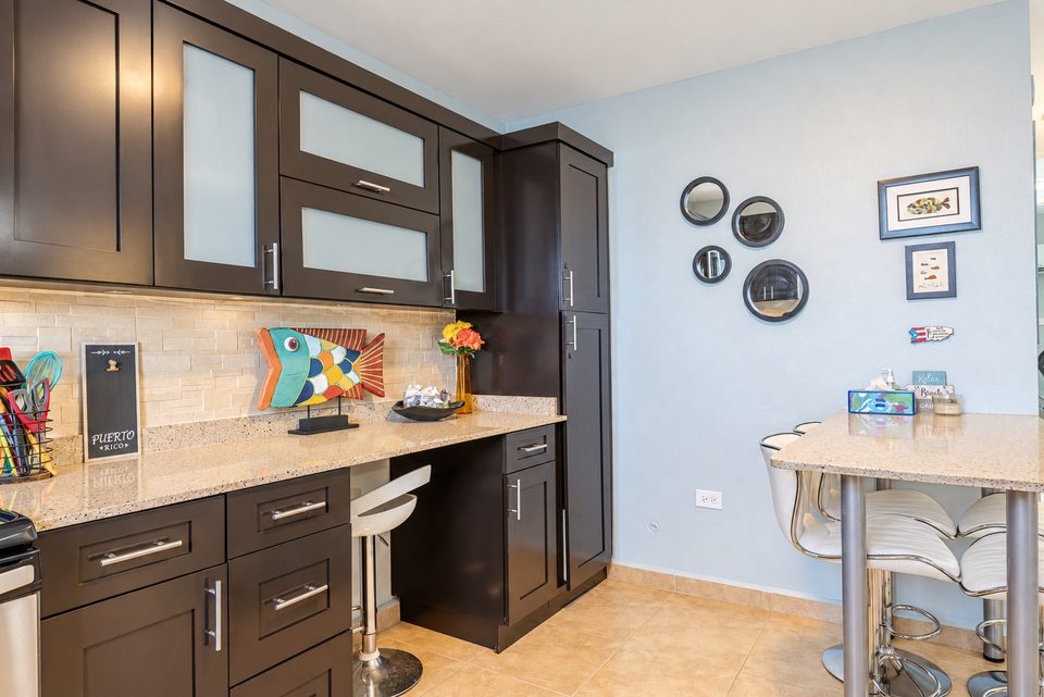 The breakfast bar with a white stool, dark cabinets, stone counters, fish art and framed prints on the pale blue wall