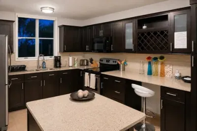 Cocina con gabinetes oscuros, topes de piedra, estufa eléctrica, microondas, cafetera y tostadora junto a la ventana, y barra con una banqueta blanca