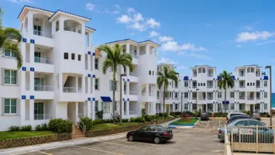Edificios blancos del condominio de tres pisos, con detalles azules, palmas y estacionamiento