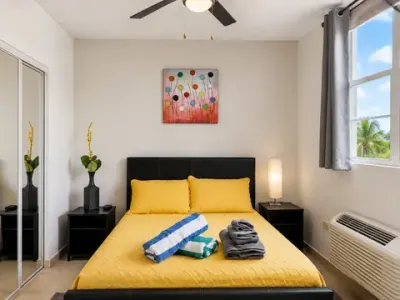 Primera habitación: cama queen con colcha amarilla y toallas dobladas, lámparas de mesa de noche y abanico de techo