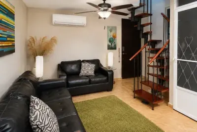 Sala con sofá y sofá de dos plazas en cuero negro, alfombra verde, escalera de caracol roja y negra al altillo, abanico de techo y puerta de entrada
