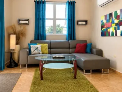 Sala con sofá seccional gris, mesa de centro de cristal, alfombra verde, aire acondicionado tipo consola y ventanas luminosas