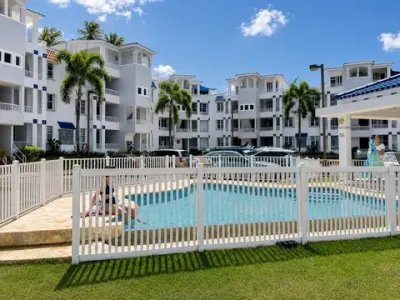 Piscina compartida dentro de una verja blanca, con los edificios del condominio y palmas detrás