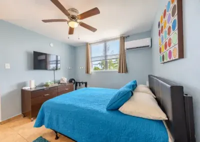 Segunda habitación: cama con colcha azul, televisor en la pared sobre el gavetero, abanico de techo, aire acondicionado tipo consola y ventana