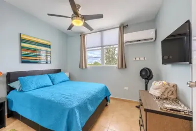 Segunda habitación desde la puerta: cama azul bajo la ventana, arte de rayas en la pared y televisor a la derecha