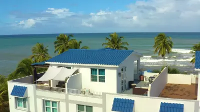 La azotea del penthouse vista desde arriba: techo de tejas azules, paredes blancas, sala exterior con sombra, terraza, palmas y el Atlántico al fondo