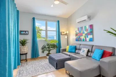 Sofá seccional gris bajo cortinas turquesa, ventana con una vista al mar, lámpara de piso y planta